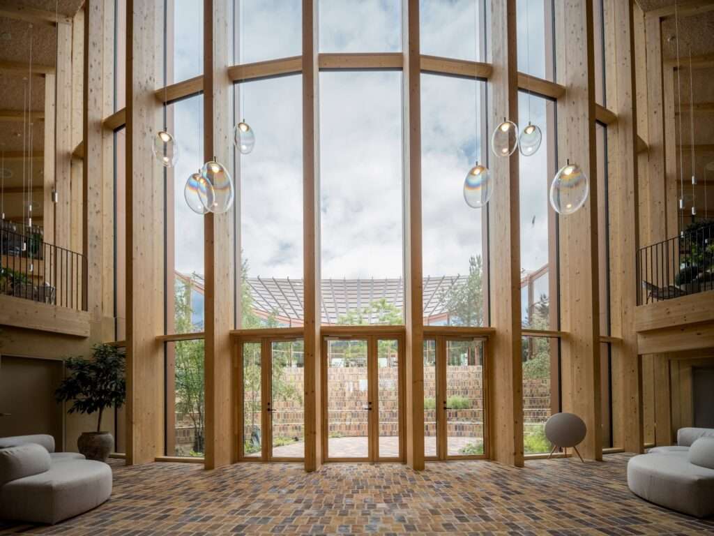 Double-height reception area of Dymak HQ with high timber columns, hanging glass lamps, and glass doors opening to a central courtyard.