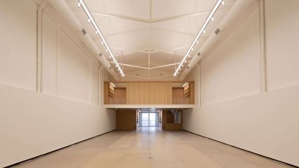 Wide symmetrical interior photo of the Larivière Foundation showing the gabled roof with white industrial trusses, white walls, polished concrete floors, and a central oak mezzanine bridge.