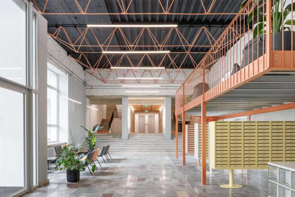 Spacious residential lobby with double-height ceiling, salmon-pink steel structural frame, exposed black corrugated ceiling, and concrete stairs.