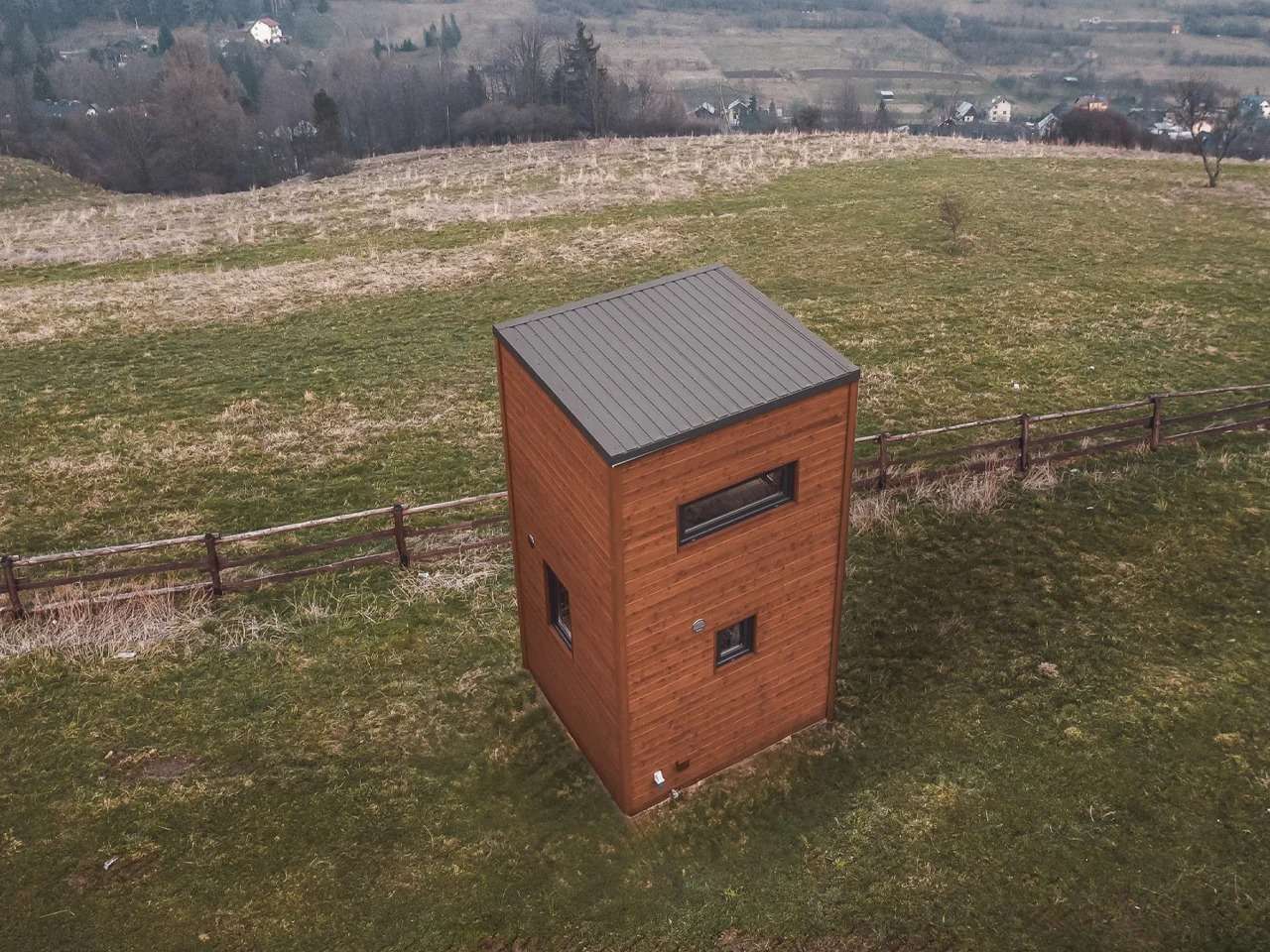 Aerial drone view of La Ruche micro-home, a compact vertical wooden cabin situated on a grassy hill with a rustic fence backdrop.