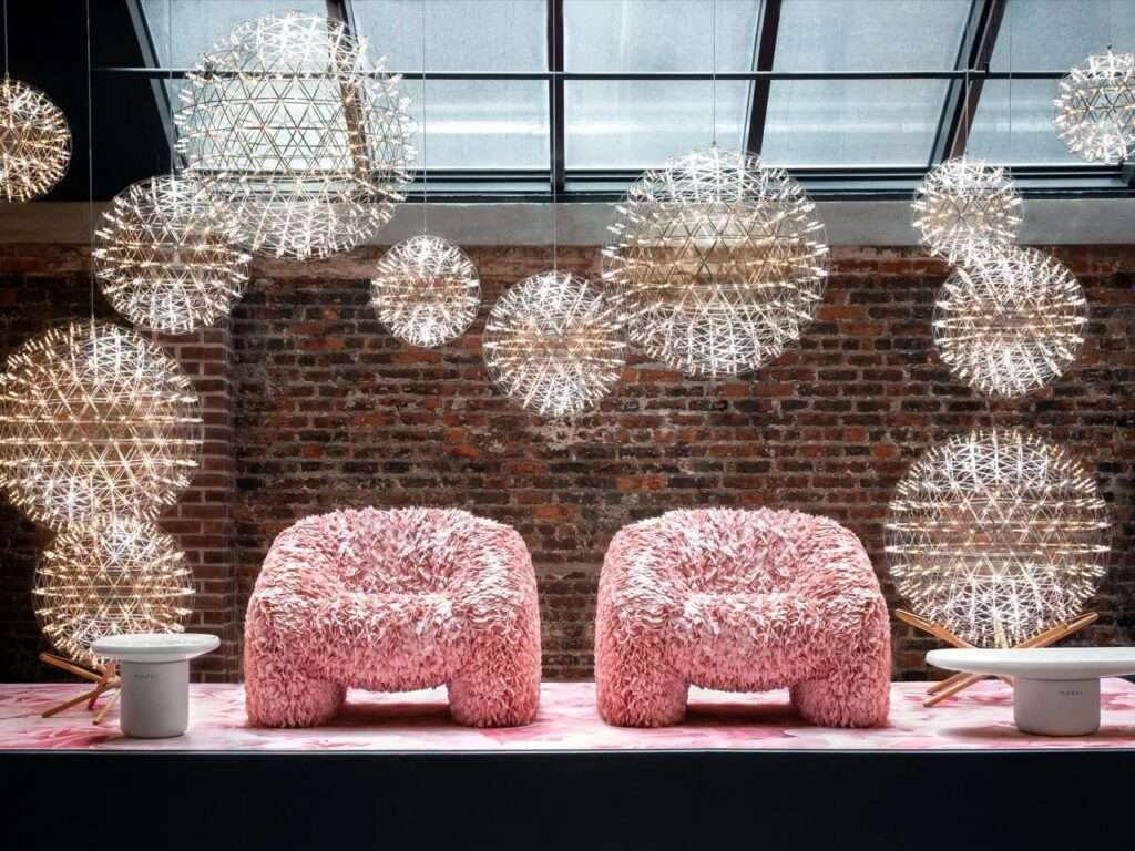 Two textured fluffy pink armchairs set against a rustic brick wall under a dense cluster of glowing spherical wire mesh pendant lights by Moooi.