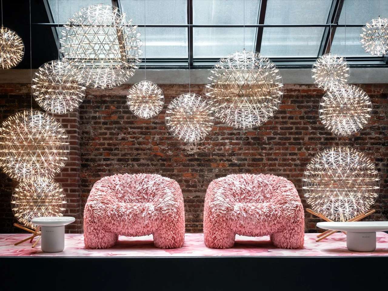 Two textured fluffy pink armchairs set against a rustic brick wall under a dense cluster of glowing spherical wire mesh pendant lights by Moooi.