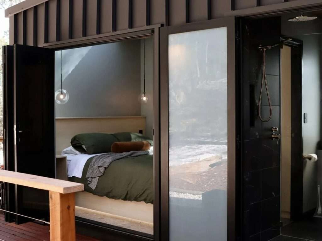 View through large, open black-framed glass doors from a wooden deck, revealing a bedroom with olive green bedding and a black-tiled bathroom with a brass shower.