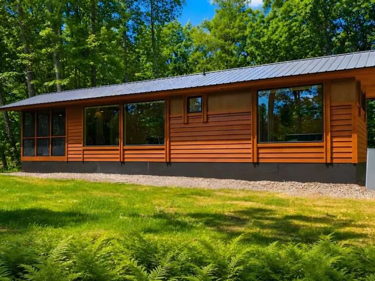 Exterior view of the Shoreline tiny home showing its elongated wooden structure, Scandinavian chalet-inspired horizontal wood siding, dark metal roof, and large panoramic windows facing a grassy green lawn.