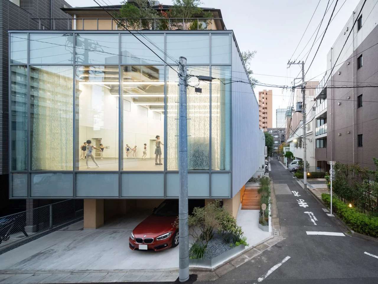 Exterior daytime view of the Tokyo Ballet Studio in Komagome showing dancers rehearsing behind a large glass facade elevated above ground level.