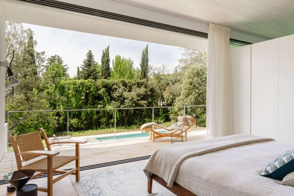 View from a modern master bedroom looking out onto a wooden deck balcony with a classic woven chair, a lounge chair, and a glass railing, overlooking a dense green hedge and pool.