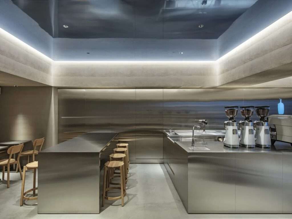 Seating area with wooden stools, stainless steel communal table, and reflective blue-tinted ceiling cavity at Blue Bottle Coffee Harajuku.