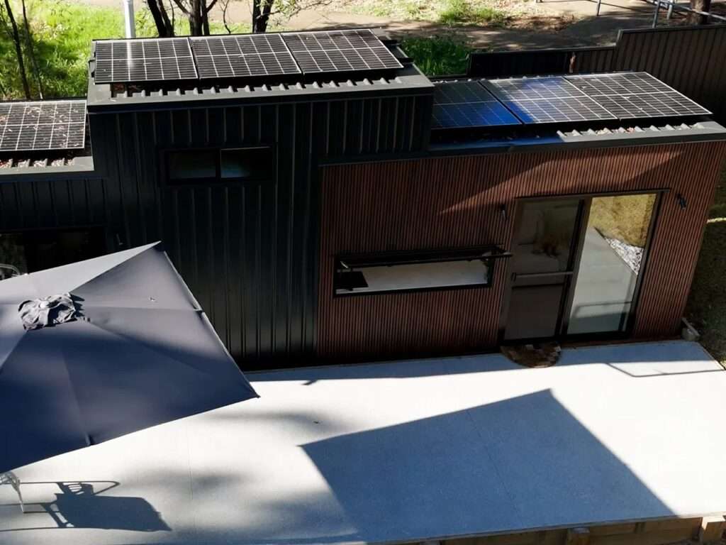 Aerial bird's-eye view of the Burley 9.6 modular home showing rooftop integrated solar panels on its stepped roofline and an outdoor concrete patio with a large umbrella.