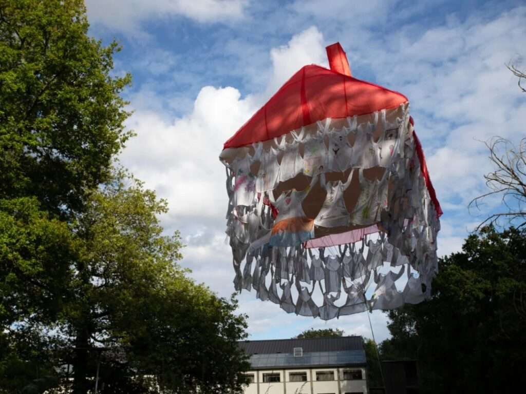 A low-angle close-up view of a floating house installation featuring a vibrant red fabric roof and rows of white upcycled t-shirts suspended underneath, set against a bright blue sky with scattered clouds and treetops.