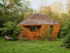 Sustainable wood shingle cabin EcoNeo tiny house exterior surrounded by lush trees and green grass.