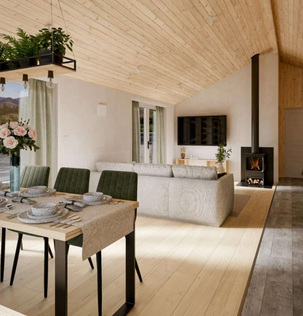 Interior view of the Evergreen XL living room looking towards a modern black wood-burning stove and fireplace, with a grey sofa and a glimpse of the outdoor scenery through large windows.