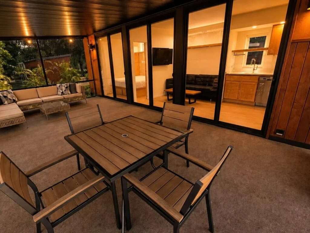 View from a wooden deck looking inside the N2 House through large glass doors, revealing the open-plan living room, kitchen, and a separate bedroom.