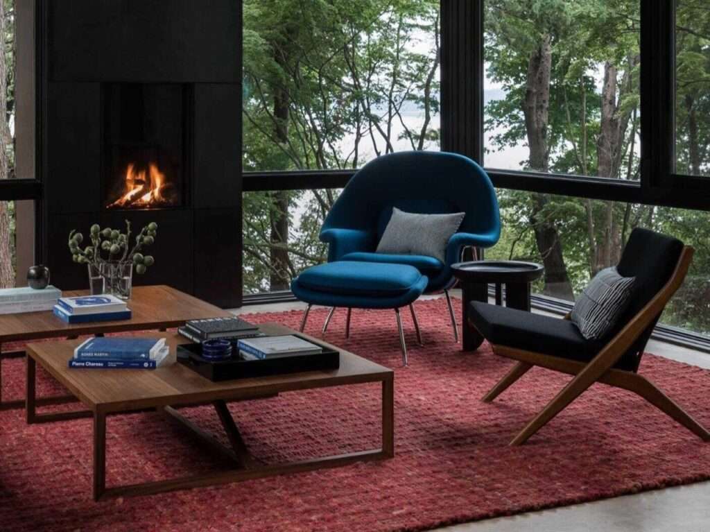 Architectural interior detail of a modern living room featuring a blue womb chair, wooden coffee tables, and a red textured rug next to a black fireplace.