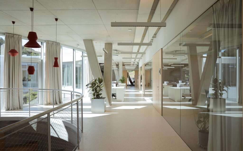 Bright open-plan office corridor featuring V-shaped concrete columns, glass partitions, red hanging pendant lights, and smooth light-colored floor finishes.