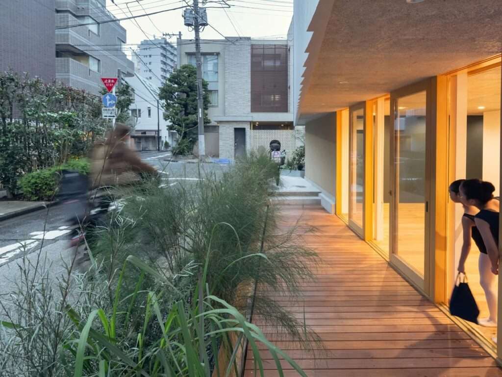 Young ballet dancers looking out from the ground-floor entrance across a wooden deck framed by lush greenery along the street.