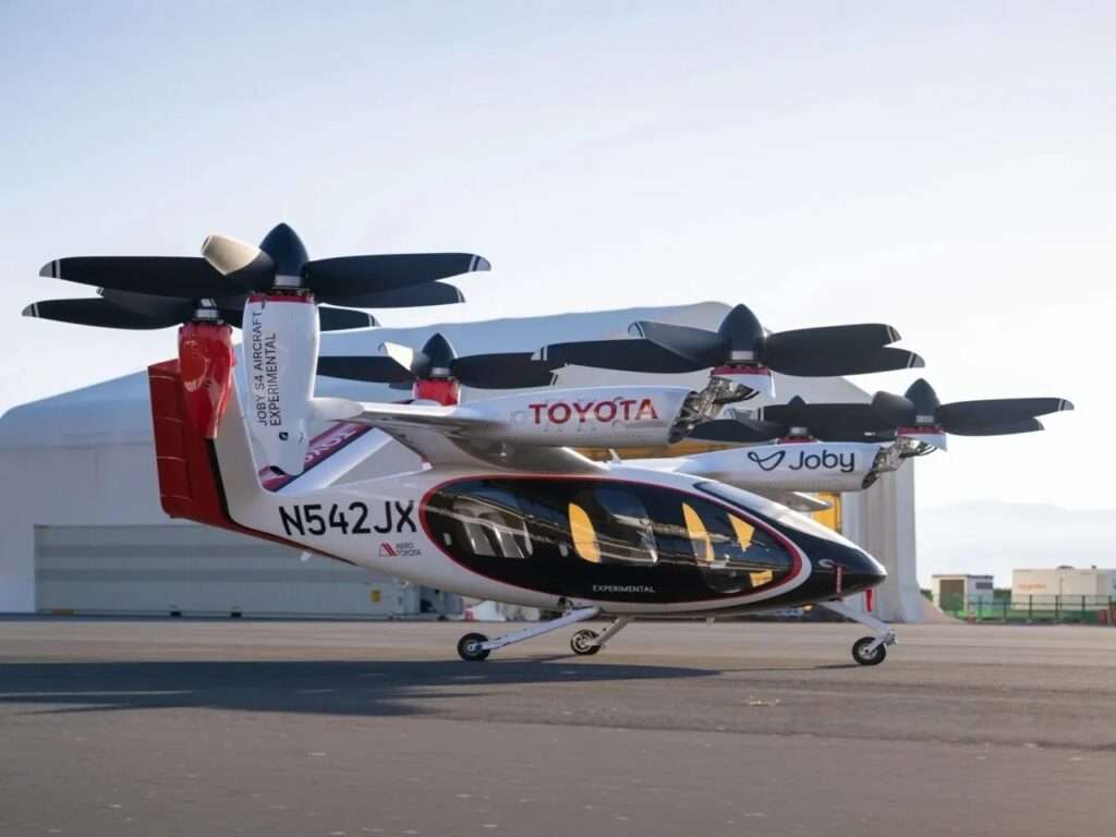 A close-up, elevated dynamic studio illustration of the Joby S4 eVTOL aircraft (N542JX), capturing the detail of the six distinctive, six-blade, tiltable prop-rotors. This diagrammatic view highlights the complex aerodynamics and is part of a larger overview of the Joby Toyota manufacturing partnership.