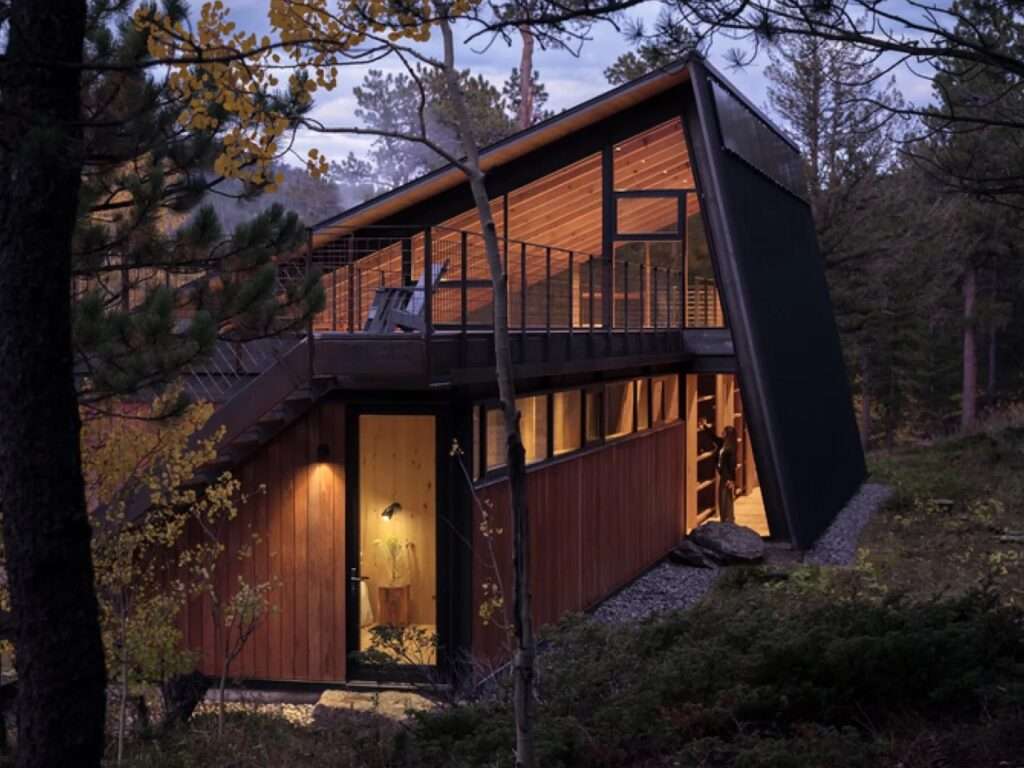 Twilight photograph of a contemporary two-story mountain cabin with illuminated glass facades, warm wood interiors, a steep metal roof structure, and an upper-level terrace nestled in a pine forest.