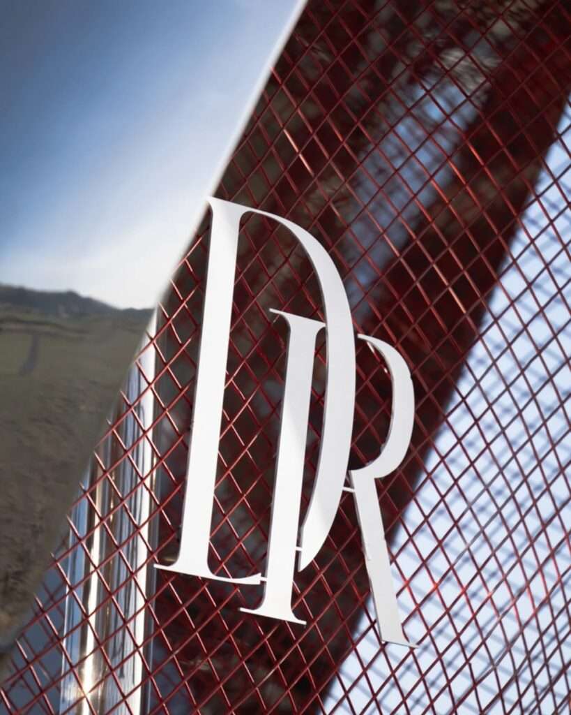 Macro close-up of a white reflective DR logo mounted on a red wire mesh structure beneath a polished chrome architectural frame.