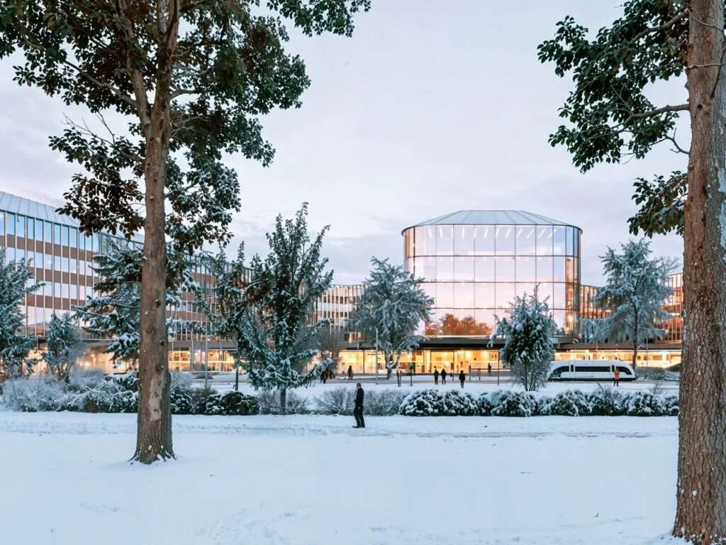The glowing glass facade of a modern hospital building in Brescia seen through snow-covered evergreen trees during winter.