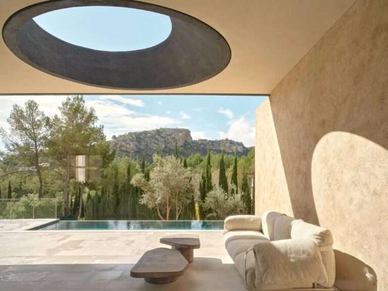 A view from an interior living area out to a pool and mountains, showing a circular oculus opening in the concrete ceiling casting a shadow on the wall.