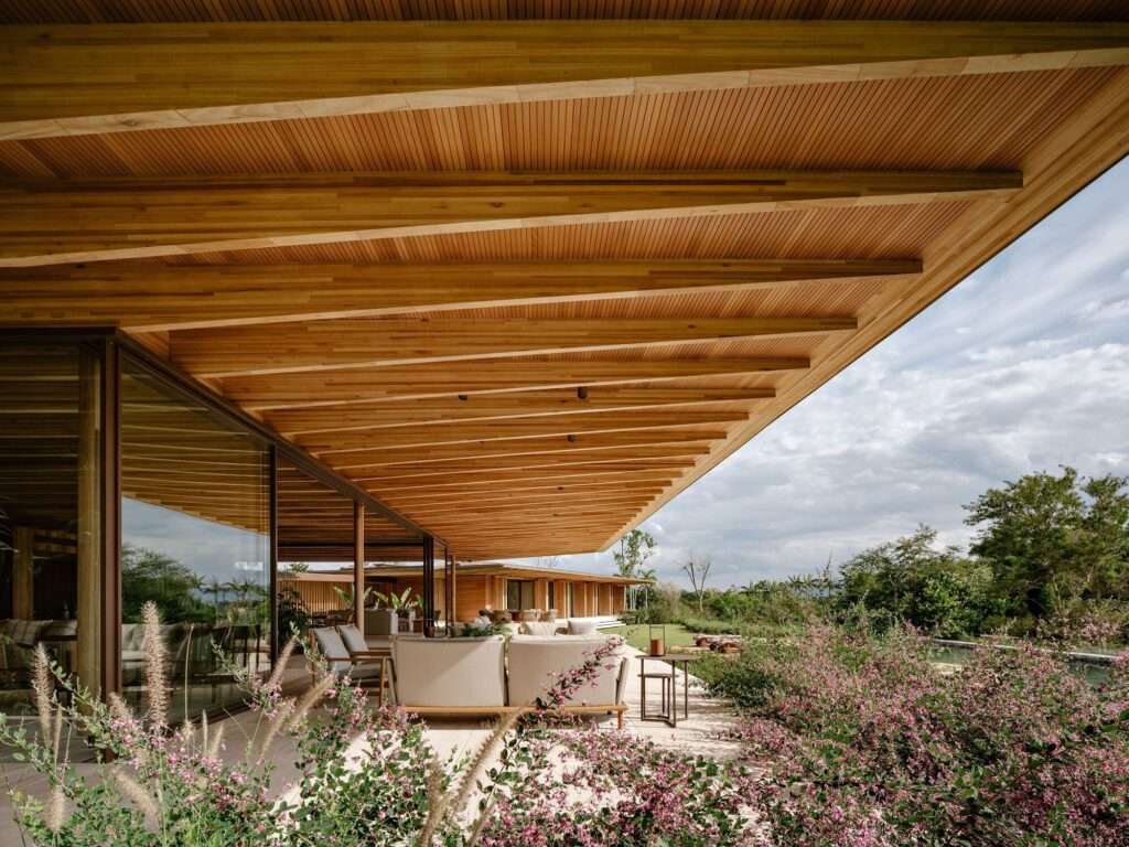 View from the veranda of AMS House looking out to the landscape, showing the large overhanging laminated timber roof with exposed beams and outdoor lounge seating.
