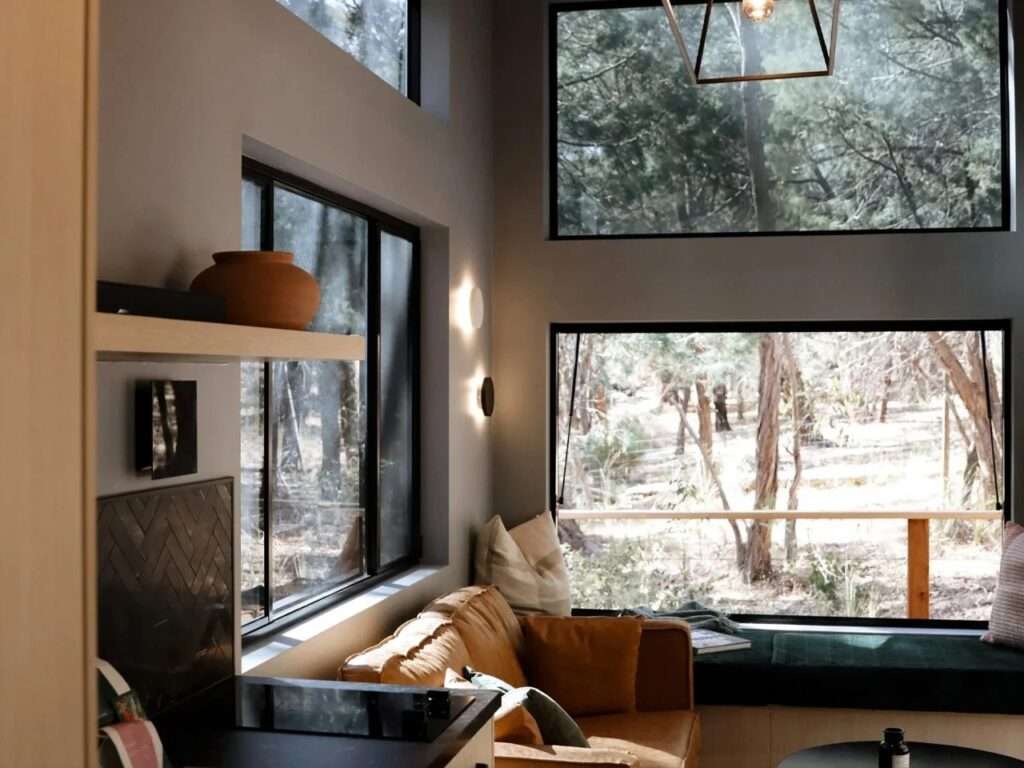 Interior of a small cabin living room with high windows framing trees, a tan leather sofa, an olive green window seat, and a geometric pendant light.