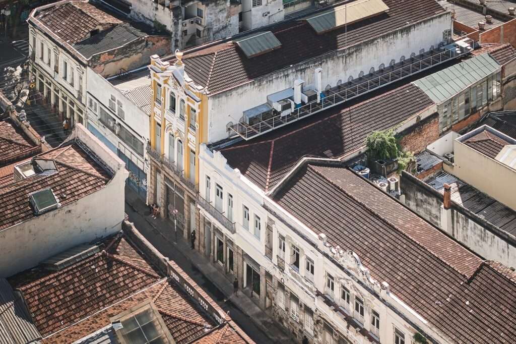 Aerial photograph of the Fábrica Bhering adaptive reuse project in Rio de Janeiro, Brazil. A central building with a prominent yellow facade and terracotta tiled roof is surrounded by a dense urban fabric of older structures. Photographed by Rafael Salim.