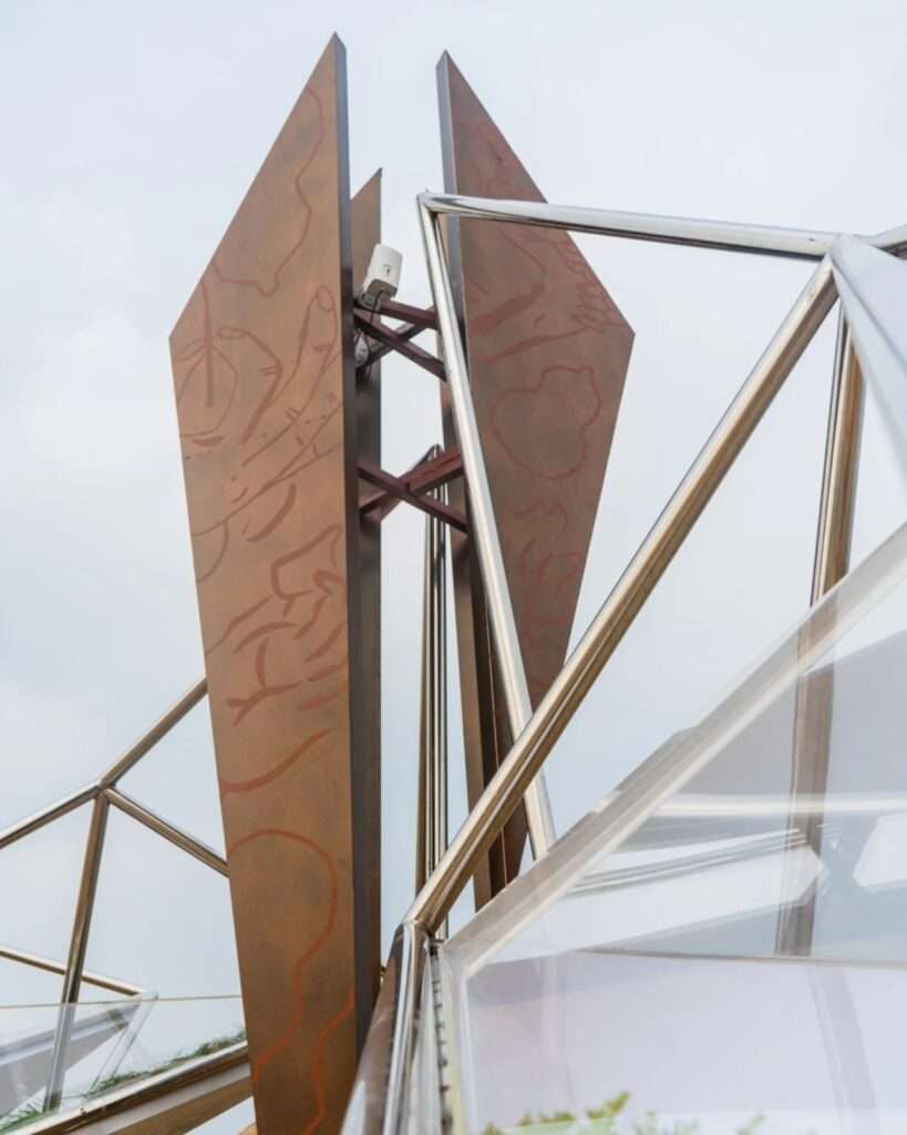 Low-angle shot looking up at etched metallic panels, structural steel joints, and transparent acrylic casing.