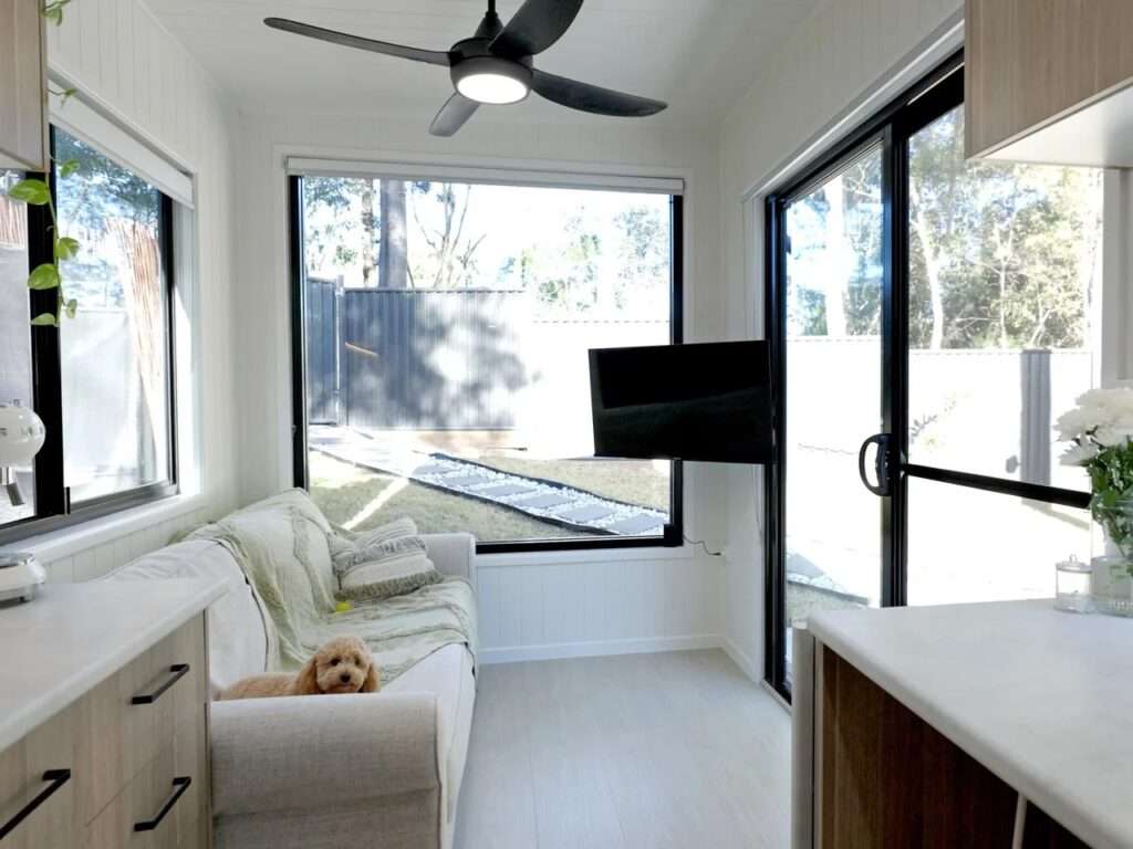 Bright compact living room interior of Burley 9.6 featuring a white fabric sofa, large panoramic glass windows, a black ceiling fan, and a wall-mounted rotating TV bracket.