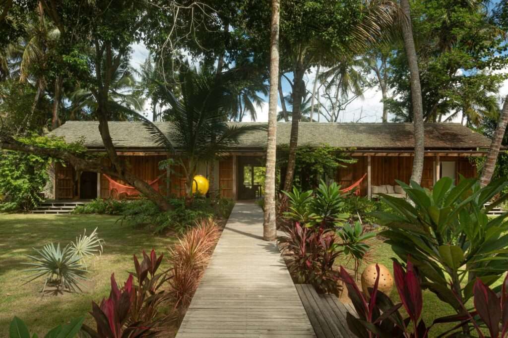 Wooden elevated walkway leading through colorful tropical plants toward a single-story wooden residential pavilion.