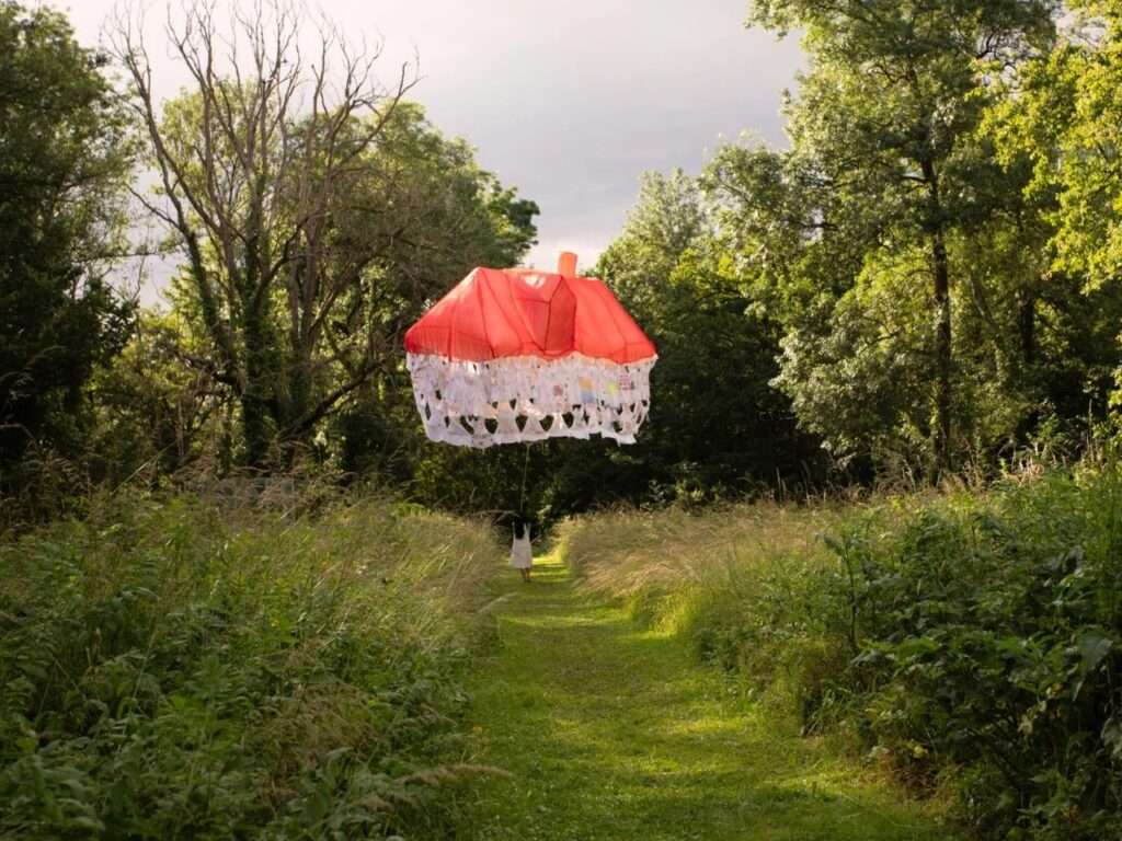 A woman walking along a narrow grassy path between lush green bushes, pulling a floating red and white house-shaped installation behind her under a soft, overcast sky.