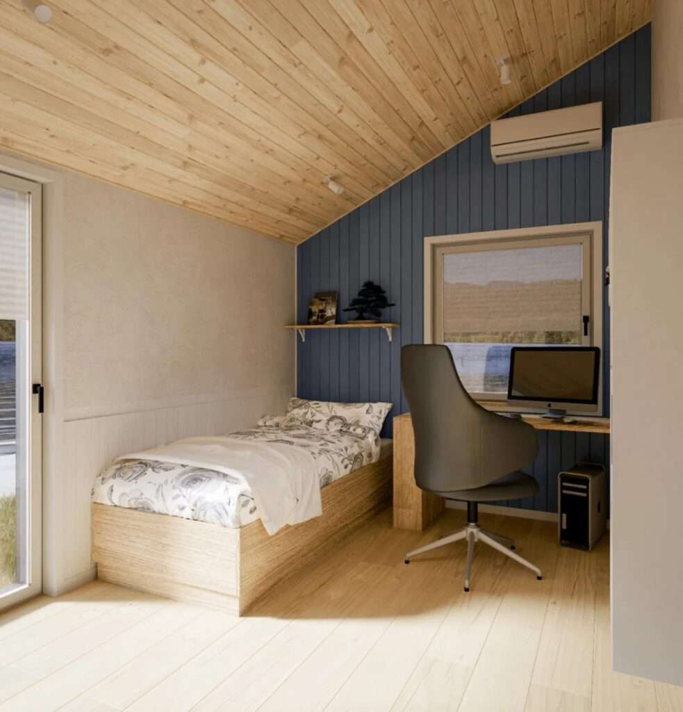 A view of the secondary bedroom in the Evergreen XL, set up as a combined workspace with a single bed next to a blue accent wall, a desk with a computer, and a glass door leading outside.