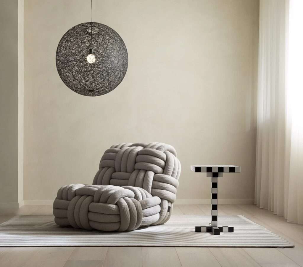 A sculptural grey accent chair crafted from thick intertwined woven ropes, positioned under a black mesh spherical pendant light over a striped rug.