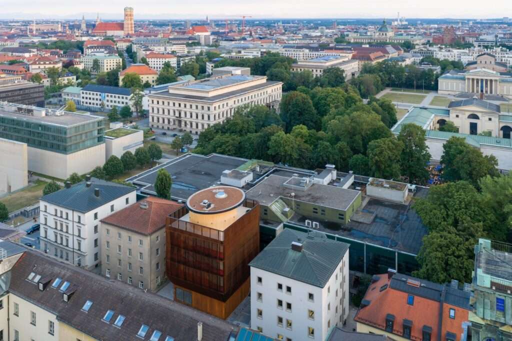 Aerial view of Munich's cityscape highlighting the timber rooftop structure of the Kinderoase an der TUM neatly tucked between historic academic buildings.