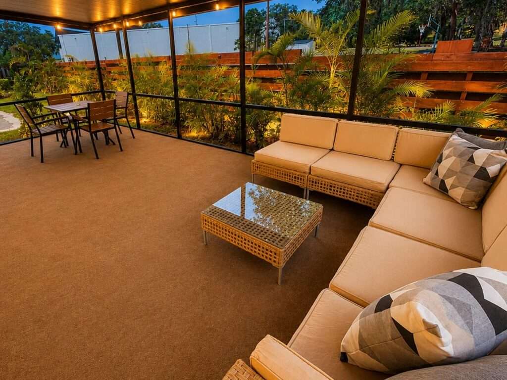 Inside a large, covered wooden deck with warm string lights, featuring a wicker sectional sofa and a glass coffee table, with palm trees visible outside.