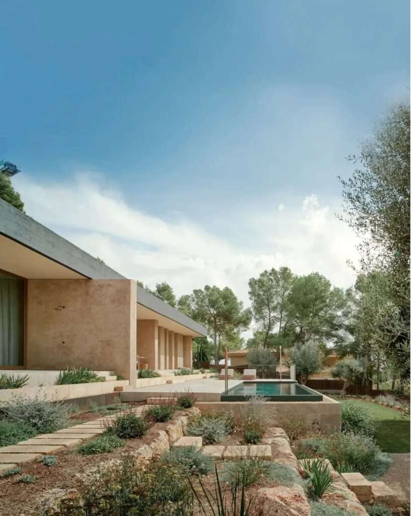 Exterior wide angle view of Casa Óculo showing its horizontal concrete roof, integrated stepped landscape with Mediterranean plants, and a swimming pool.