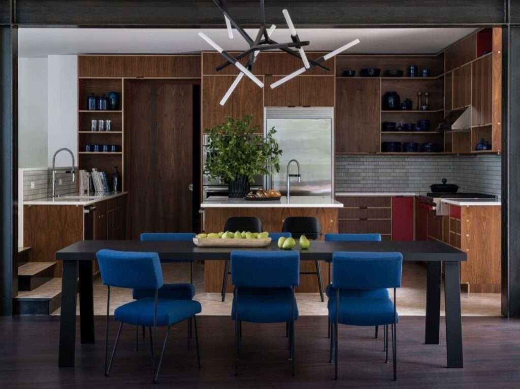 Modern open-plan dining table with bright blue chairs set against walnut wood kitchen cabinetry and a sculptural black chandelier.
