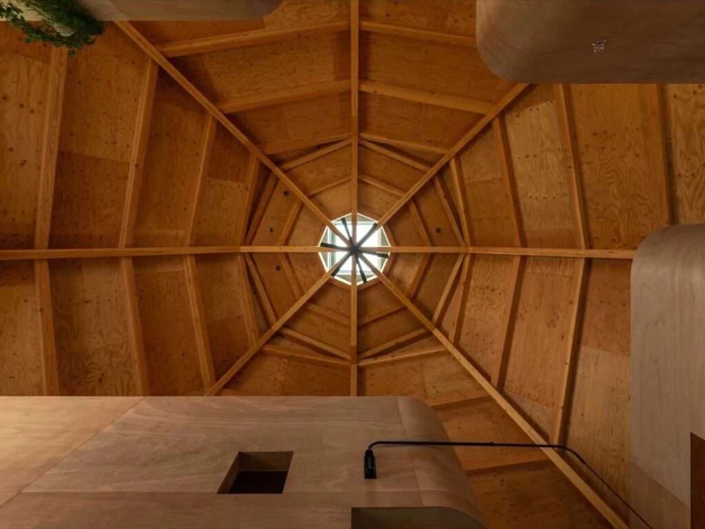 Directly upward looking shot at a radial octagonal timber roof structure converging around a central skylight with a black ceiling fan.