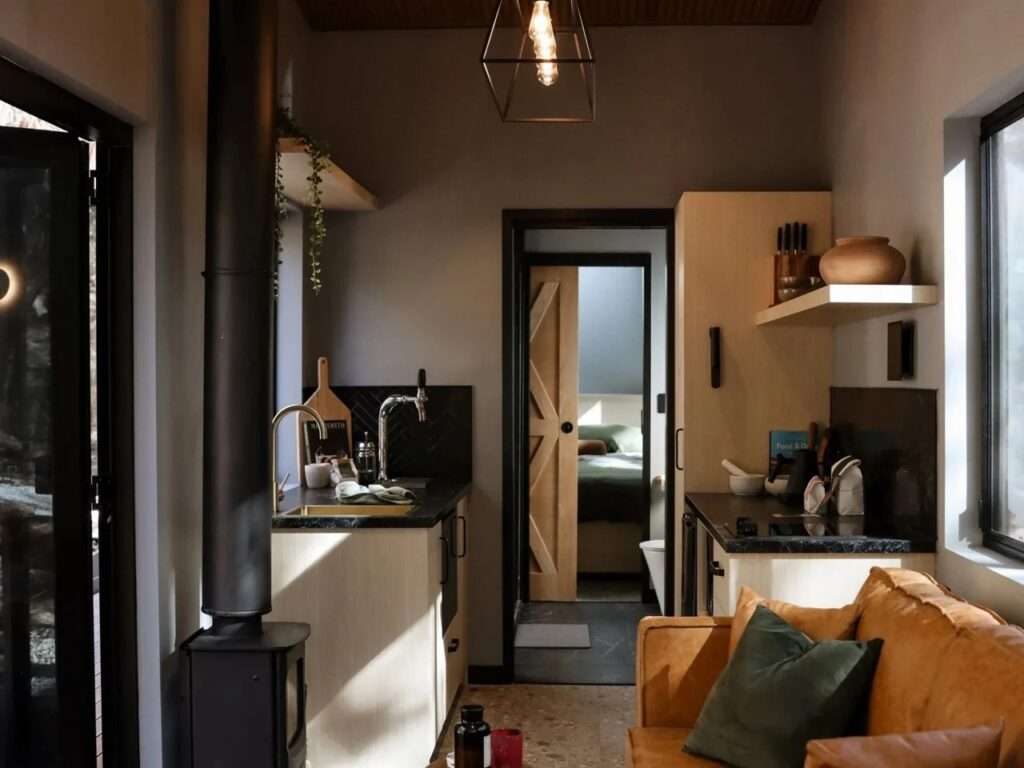 Compact modern cabin kitchen featuring light wood cabinets, dark stone counters, a black wood-burning stove, copper fixtures, and a wooden sliding barn door leading to a bedroom.