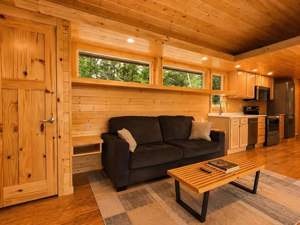 Warm all-wood interior of a living room inside a tiny house featuring a dark grey sofa, a slatted wooden coffee table, and an open layout leading to a modern kitchen.