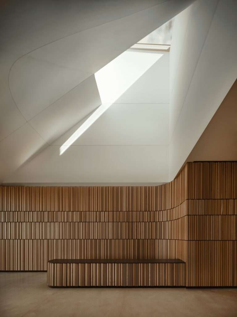 Minimalist interior space featuring curved wooden slat walls and a dramatic skylight casting a bright beam of natural light.