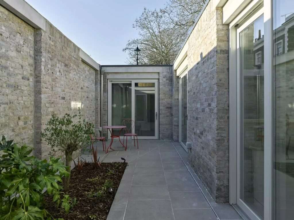 Internal courtyard garden with grey porcelain tile paving, reclaimed brick walls, and large sliding glass doors.