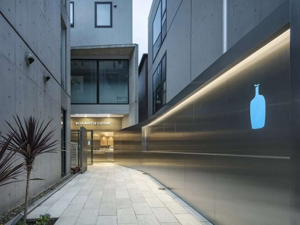 Exterior passageway leading to Blue Bottle Coffee Harajuku with a long curved stainless steel wall and glowing blue logo.