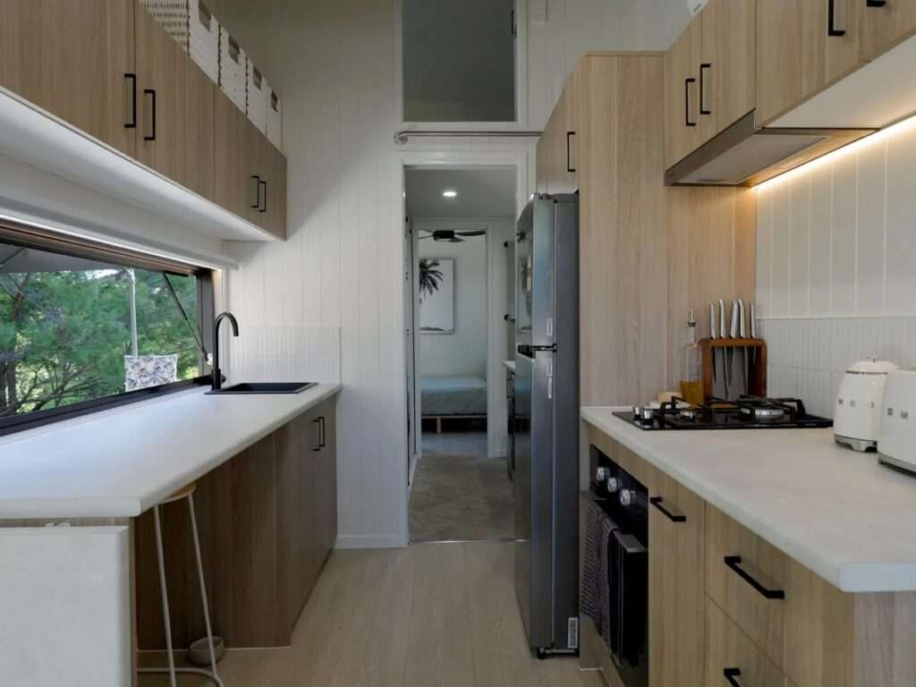 Linear corridor kitchen inside the Burley 9.6 tiny home with light oak cabinetry, white VJ groove walls, stainless steel refrigerator, and a large pass-through window.