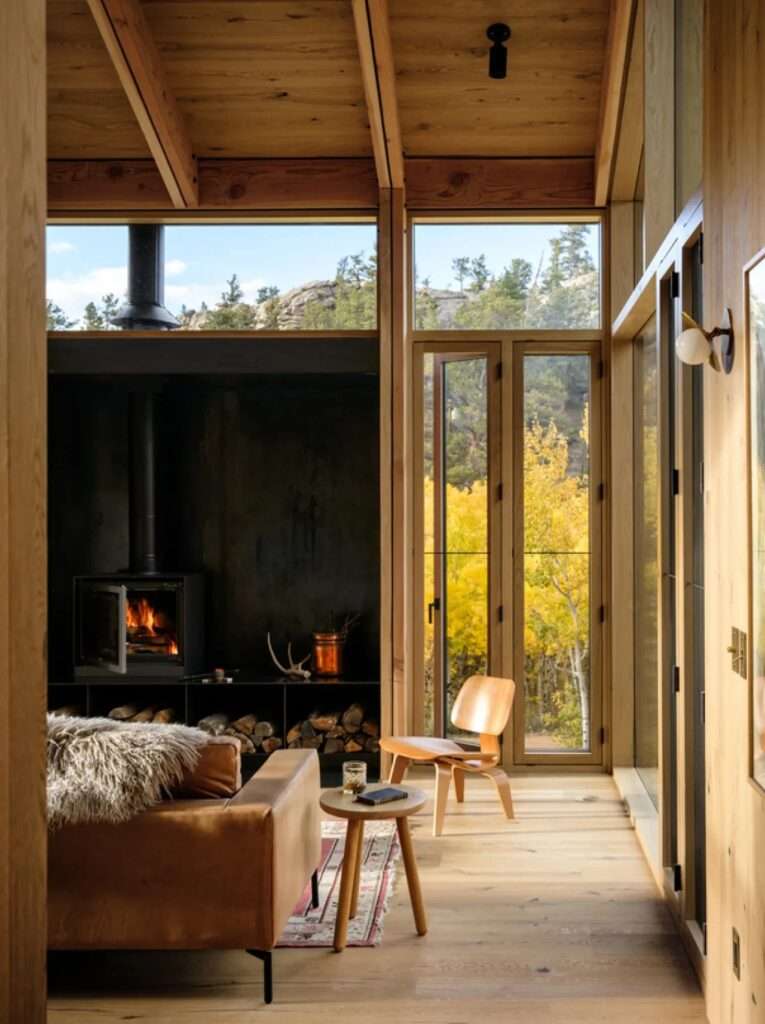 Minimalist cabin interior featuring rustic oak flooring and walls, a leather sofa, a mid-century modern wooden chair, and a central black wood-burning fireplace alongside floor-to-ceiling windows.