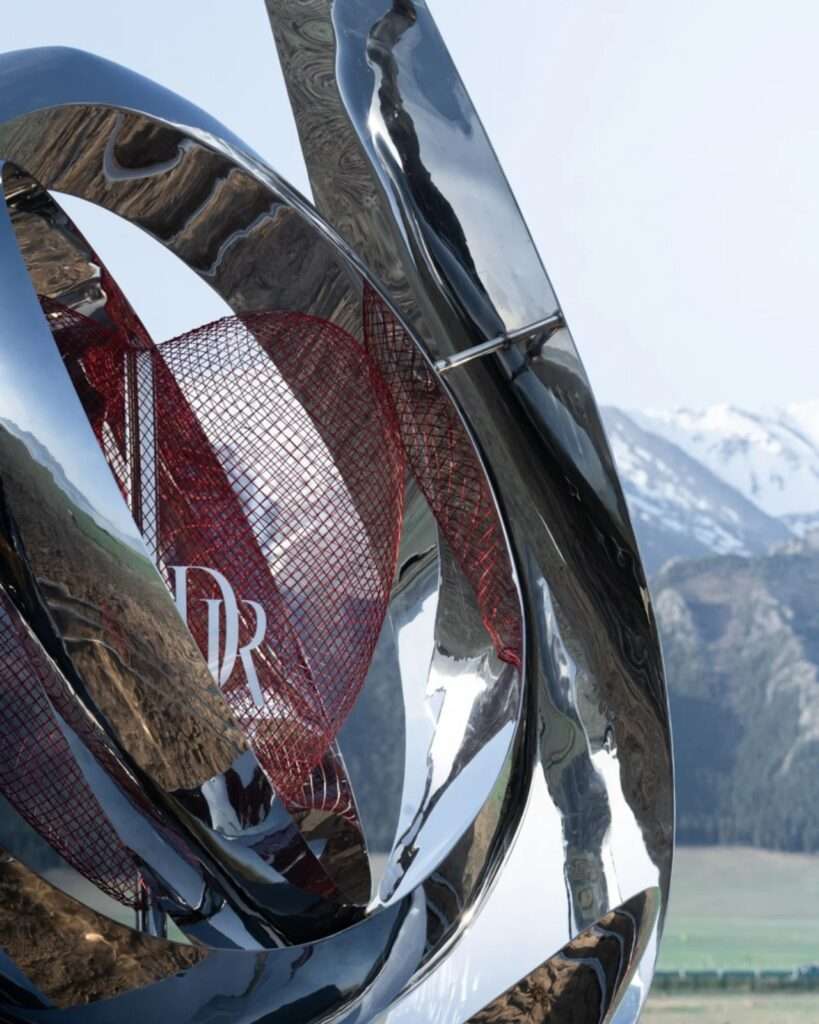 A close-up view of the interlocking curved metal bands reflecting the surrounding green hills and snow-capped mountains, with the red mesh core visible inside.