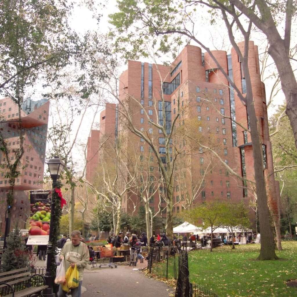 Urban park pathway with mature green trees leading toward the redeveloped brick residential high-rises under a clear sky.