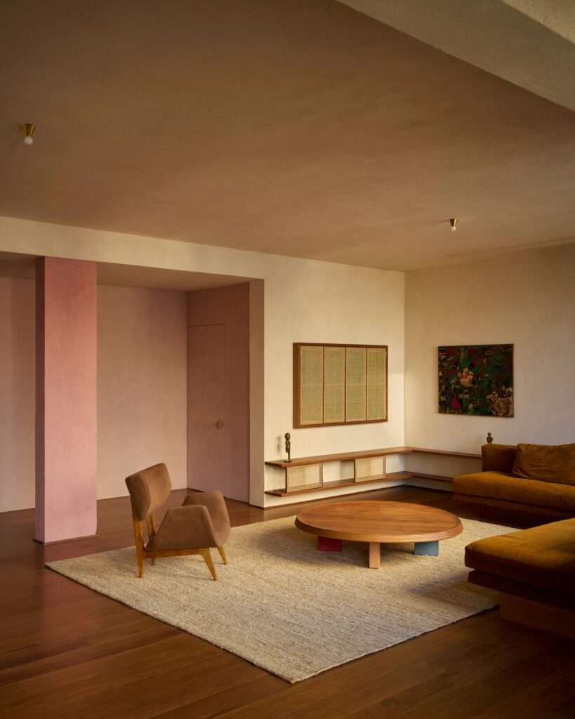 Wide shot of the main living room showing the pink structural column, a bouclé armchair, round wooden coffee table on a jute rug, custom built-in wooden shelving, and a mustard-yellow sofa.