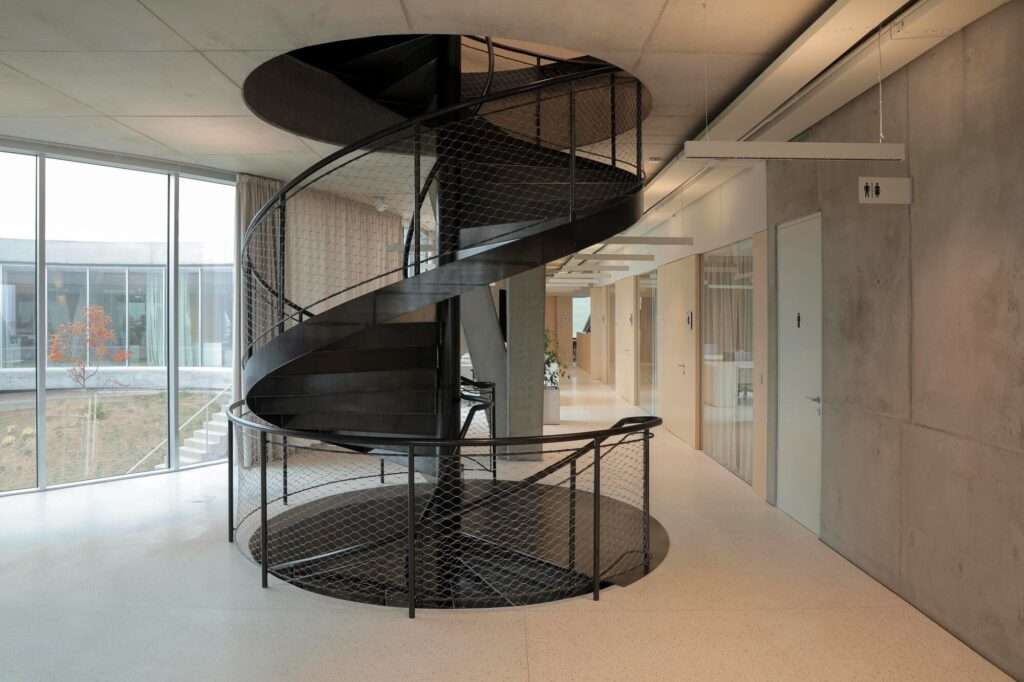 Sleek black metal spiral staircase connecting the upper office floor to the lower courtyard and multifunctional spaces, positioned next to a glass curtain wall.