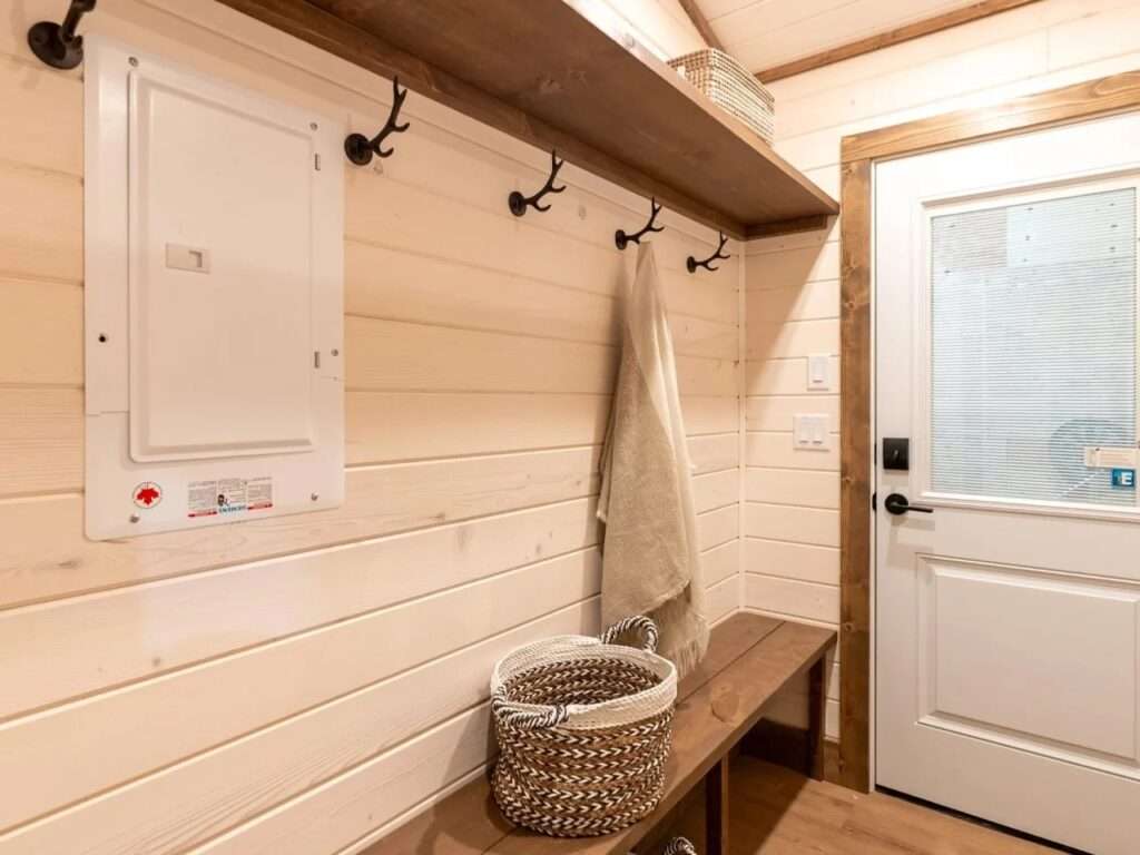 Cozy mudroom in a tiny house featuring a dark wood bench, antler-style coat hooks, an overhead storage shelf, and a white entry door.
